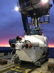 A white submersible vehicle is secured on the deck of a ship at sunset, its onboard lights illuminating the vessel and ocean in the background, evoking a sense of the sublime amid the tranquil seascape.